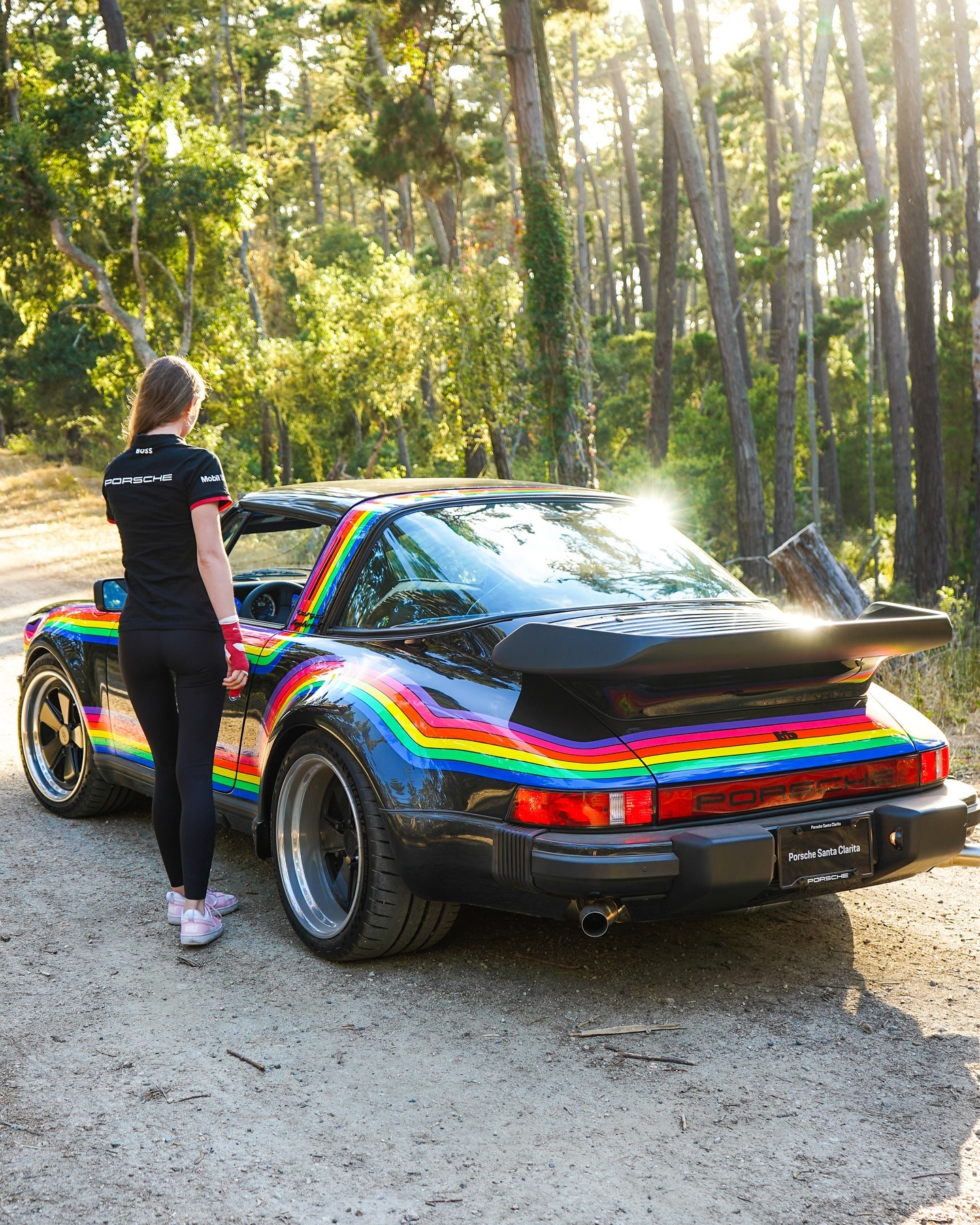Kaytea standing beside a colorful Porsche on a wooded road